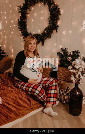 Schwangere Frau sitzt im Schlafanzug mit auf dem Bett im Schlafzimmer. Weihnachtsmorgen. Das Interieur des neuen Jahres. Valentinstag-Feier Stockfoto