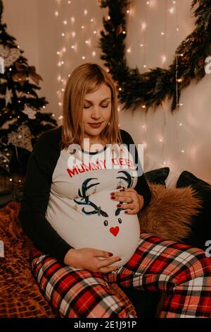 Schwangere Frau sitzt im Schlafanzug mit auf dem Bett im Schlafzimmer. Weihnachtsmorgen. Das Interieur des neuen Jahres. Valentinstag-Feier Stockfoto