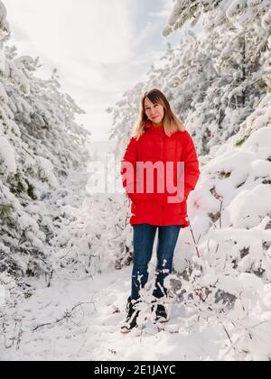 Junge Frau in roter Jacke zwischen verschneiten Bäumen im Winterwald. Reisende Frau mit Schneebäumen in sonnigen Tag Stockfoto