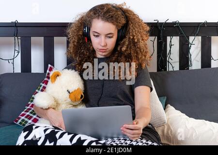 Ein junger lockiger Teenager mit einem Teddybär sitzt darauf Das Bett und schaut einen Film auf ihrem Laptop Stockfoto