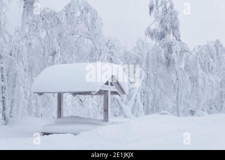 Verschneite hölzerne Gartenlaube mit Picknicktisch in einer Schneeverwehung nach Ein starker Schneefall in einem frostigen Winterpark Stockfoto