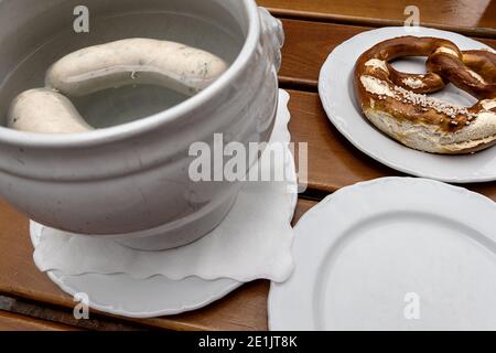 Zwei bayerische Weißwürste in heißem Wasser, eine Brezel auf einem Teller und ein leerer Teller Stockfoto