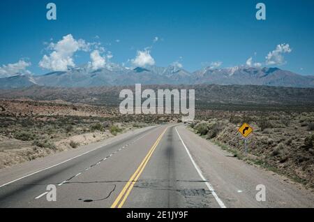 Leere Asphaltstraße mit Kurven, gelber Trennlinie und Verkehrspfeilzeichen in einem Wüstental in den Anden, Provinz Mendoza, Argentinien Stockfoto