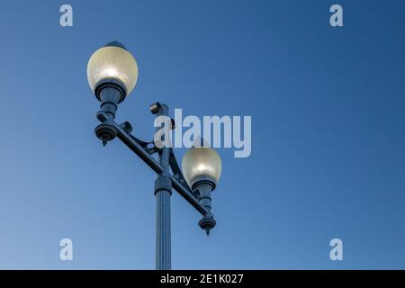 Ein Stadtlampenposten ist aus einem niedrigen Winkel gegen einen gradienten blauen Himmel zu sehen. Die Leuchten sind in einem nebligen Glasgehäuse auf einem blauen Metallständer beleuchtet. Stockfoto