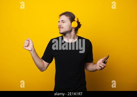 Langhaariger Mann mit Bart tanzt mit Kopfhörern und Halten Sie ein Handy an einer gelben Studiowand mit einem Schwarzes T-Shirt Stockfoto