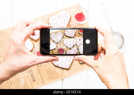 Hände mit dem Telefon Nahaufnahme Bilder hausgemachte Cookies für valentinstag Tag mit Herzen auf einem hellen Hintergrund bestreut Stockfoto