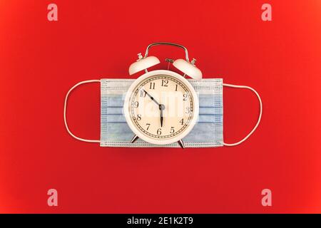 COVID-19 Konzept, neue Morgenkomposition, Maske vor dem Ausgehen tragen, Wecker und Zeitkomposition auf rotem Hintergrund, Foto Stockfoto