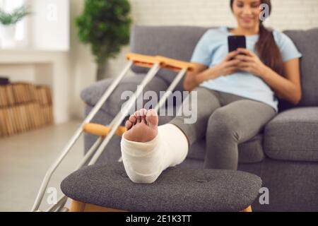 Frau mit gebrochenem Bein in Gips gegossen sitzt auf der Couch Zu Hause mit Handy in den Händen Stockfoto
