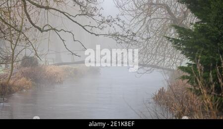 Holzbrücke über kleinen Fluss durch einen schweren Nebel gesehen An einem Wintertag Stockfoto