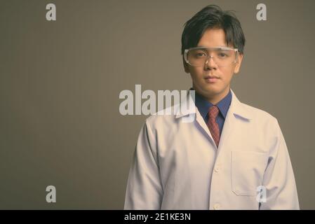 Junge gut aussehende asiatische Mann Arzt trägt Schutzbrille gegen grau Hintergrund Stockfoto