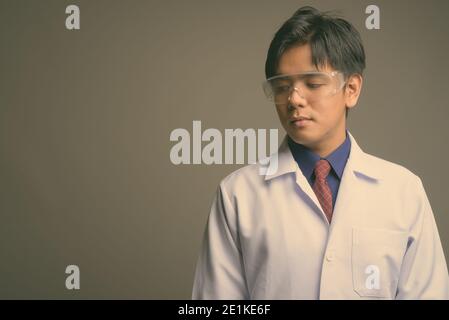 Junge gut aussehende asiatische Mann Arzt trägt Schutzbrille gegen grau Hintergrund Stockfoto