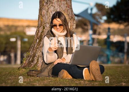 Foto von glücklichen kaukasischen Frau sitzen auf dem Gras im Freien im Park mit Laptop und Handy. Online- und Freiberufliches Arbeitskonzept Stockfoto