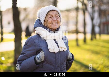 Portrait kaukasische ältere Frau mit grauen Haaren und tiefen Falten 90 Jahre alt posiert in warmer Kleidung, weiß gestrickter Schal und Hut im Park, sonniger Frost Stockfoto