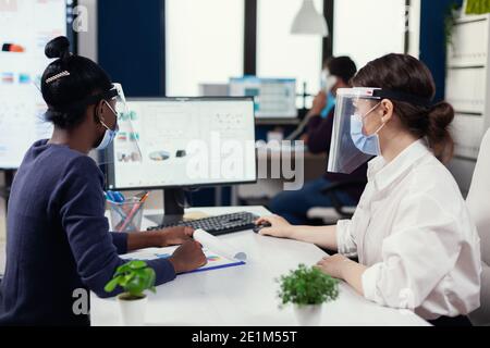 Geschäftsleute, die Computer mit Finanzgraph tragen Gesichtsmaske für covid19. Multiethnisches Team arbeitet in Unternehmen mit neuen normalen Respekt soziale Distanz während der globalen Pandemie mit Coronavirus. Stockfoto