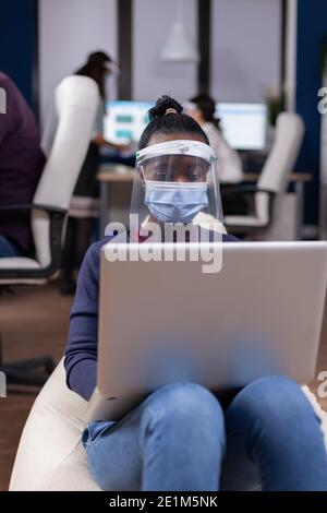 Afrikanischer Manager in comany mit Laptop trägt Gesichtsmaske gegen covid. Multiethnisches Geschäftsteam, das während der globalen Pandemie mit Coronavirus soziale Distanz respektiert. Stockfoto