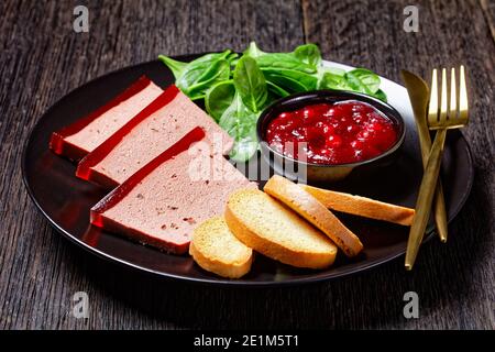 Französisches Gericht Entenleber Pastete mit Cranberry Marmelade auf einem schwarzen Teller serviert mit Cranberry-Sauce, Spinat und Brioche Croutons, mit goldenem c Stockfoto