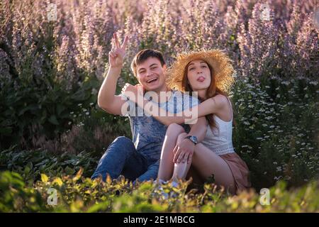 Glückliches Paar in der Liebe sitzt umarmt von blühenden Salbei Feld. Ein junger Mann mit schönen Mädchen in Strohhut haben Spaß, herumtäuschen, winken ihre ha Stockfoto