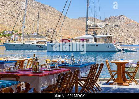 Taverna Tische und Stühle am Wasser mit Blick auf verankerte Boote in Pedi Bay, Symi Island, Dodekanes, Griechenland Stockfoto