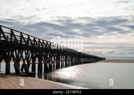 Ein langer hölzerner Steg Pier führt von einem Strand zu Ein weiterer über einem kleinen Meeresarm Stockfoto