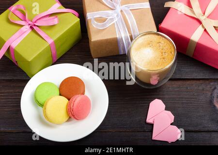 Weißer Teller mit französischen Makronen, zwei Geschenkschachteln, kleinen Papierherzen und einem Glas Latte. Ein Geschenk für einen geliebten Menschen. Stockfoto