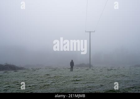 Eine geheimnisvolle Kapuzenfigur, zurück zur Kamera. Blick auf Telegrafenmasten. An einem frostigen, nebligen Wintertag. Stockfoto