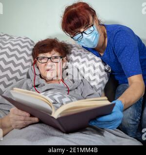 Kranke ältere Frau liegt in einem Bett und liest ein Buch, ältere Frau liest Buch mit Krankenschwester Stockfoto