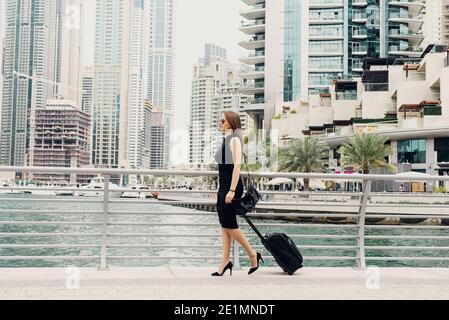 Junge moderne selbstbewusste Geschäftsfrau zieht einen Koffer in einem Dubai Marine. Einen neuen Job in einer großen Stadt zu starten. Stockfoto