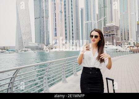 Beschäftigte junge moderne Geschäftsfrau im Anzug, die einen Koffer in einem Dubai Marine zieht. Zu ihrem Büro zu gehen und am Telefon zu sprechen. Stockfoto