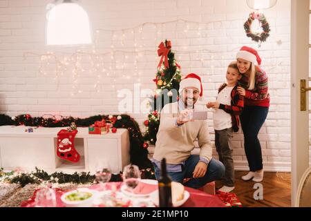 Vater fotografiert seine Familie mit dem Smartphone. Im Hintergrund Weihnachtsdekoration. Weihnachtsferien Konzept. Stockfoto