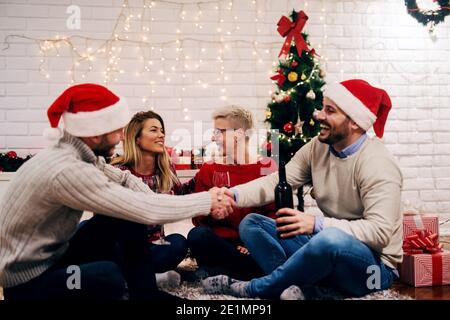 Zwei junge Paare feiern weihnachten zusammen. Sitzen auf einer Etage Spaß und Wein trinken. Bunte weihnachtsdekoration im Hintergrund. Stockfoto