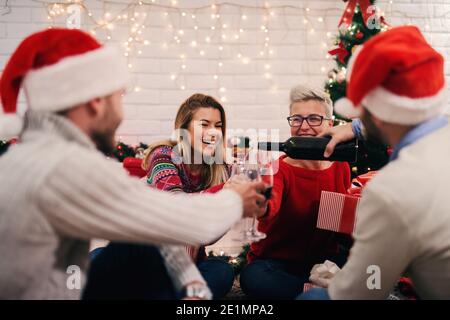 Zwei junge Paare feiern weihnachten zusammen. Sitzen auf einer Etage Spaß und Wein trinken. Bunte weihnachtsdekoration im Hintergrund. Stockfoto