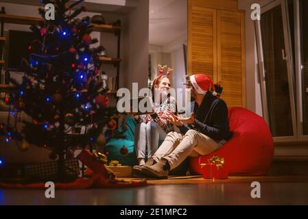 Paar mit Tablette, während sitzen in Bohnensäcken. Auf den Köpfen Rentier Stirnband und santa's Hut. Im Vordergrund Weihnachtsbaum und Geschenke. Weihnachtsurlaub Stockfoto