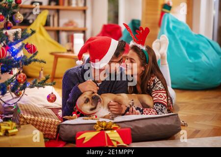 Pärchen liegen auf dem Boden und streicheln ihren Hund. Neben ihnen Weihnachtsbaum. Home Interior, Weihnachtsferien Konzept. Stockfoto