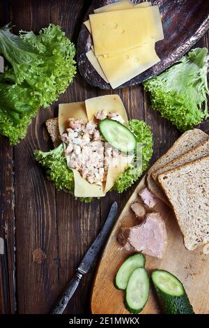 Flachlay-Nahaufnahme von frischem Salat-Sandwich auf dunkler rustikaler Holztischoberfläche mit grünen Salatblättern, Käse, geschnittenem Thunfisch, Gurken und Weizen Stockfoto