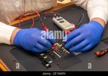 Techniker im Service-Center repariert einen LCD-Fernseher. Handschuhen messen den Widerstand auf der Rückseite des Monitors mit einem Multimeter. Selektiver Fokus, Nahaufnahme. Stockfoto