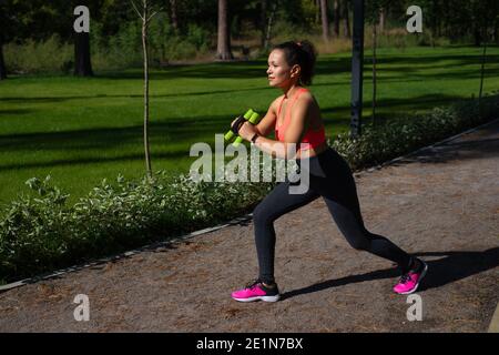 Sportliche Frau in Sportbekleidung beim Ausfallschritt mit Kurzhanteln auf einem Laufweg im Park. Fitness im Freien Stockfoto