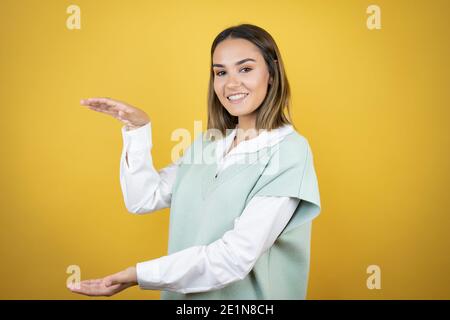 Hübsche junge Frau, die über gelben Hintergrund gestikuliert mit Händen zeigt große und große Zeichen, Maßnahme Symbol. Lächelnd. Messkonzept. Stockfoto