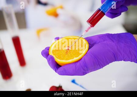 Nahaufnahme einer Hand in einem Schutzhandschuh, der Orange hält und mit einer Spritze eine Probe davon nimmt. Stockfoto
