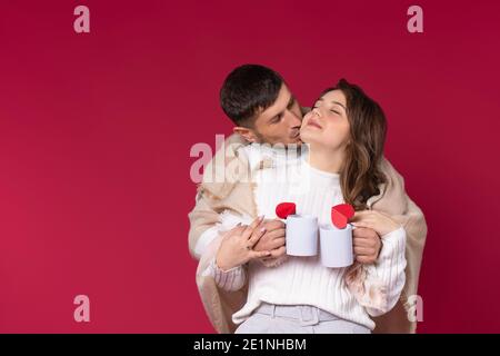 Ein liebevolles Paar in einer warmen Decke hält heißen Tee Tassen gewickelt haben eine schöne Zeit. Roter Hintergrund. Valentinstag. Stockfoto