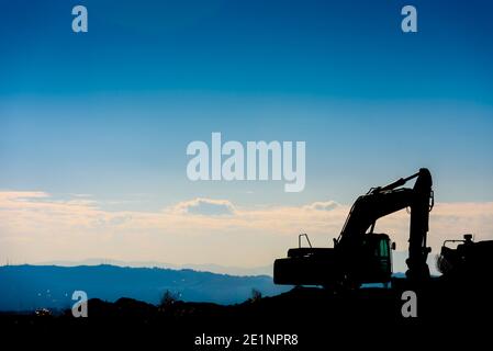 Hinterleuchtete Silhouette eines Baggers auf einem Hügel mit unfokussierten Himmel Hintergrund. Stockfoto