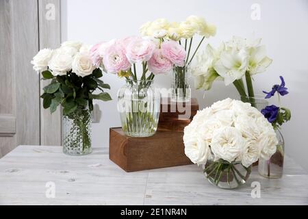 Weiße Rosen, Ranunculus, blaue Anemonen, gelbliche Butterblumen, Lilien in runden Vasen auf den Holzkisten auf dem Tisch für einen besonderen Anlass als Kitch Stockfoto