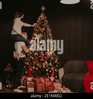 Junge Frau sitzt auf den hinteren Schultern des Mannes und schmückt einen Weihnachtsbaum, hängt Ornamente. Dekoriertes Haus für Neujahr. Weihnachten Morgen. Wohnung Stockfoto