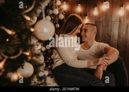 Paar in der Liebe Kerl und Mädchen umarmen küssen in der Nähe von Weihnachtsbaum in der Nähe eines Fensters auf einem hölzernen Fensterbank. Dekoriertes Haus für Neujahr. Weihnachten morgen Stockfoto