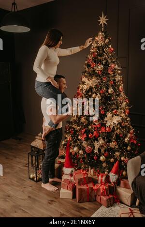 Junge Frau sitzt auf den hinteren Schultern des Mannes und schmückt einen Weihnachtsbaum, hängt Ornamente. Dekoriertes Haus für Neujahr. Weihnachten Morgen. Wohnung Stockfoto