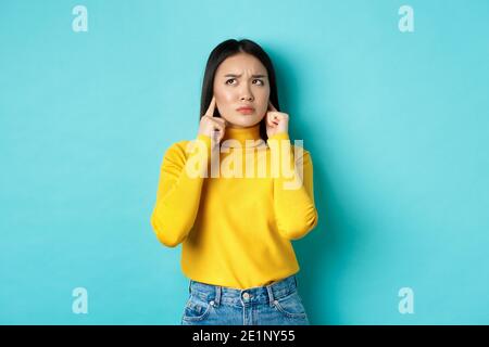 Verärgerte koreanische Mädchen beschwert sich über laute Nachbarn, Ohren mit Fingern schließen und wütend auf, stehen vor blauem Hintergrund Stockfoto