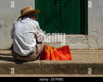 Ein Mann mit einem Hut sitzt auf einem Bürgersteig vor einer grünen Tür in einer Straße der Kolonialstadt Villa de Leyva, in der zentralen Andengebirge von Stockfoto