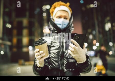 Ältere Frau hält Kaffee und Handy, trägt schützende medizinische Maske und weiße Latexhandschuhe, Spaziergänge im Freien, Beobachtung der sozialen Distanz durch Corona Stockfoto