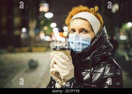 Reife kaukasische Frau in schützende medizinische Maske und Handschuhe, hält Kaffee zum Mitnehmen in den Händen in der Nacht Stadt. Ältere Frau gehen während der Quarantäne coron Stockfoto