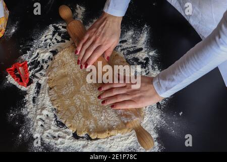 Weibliche Hände mit einem hölzernen Nudelholz bereiten Teig für Kekse auf dem Küchentisch mit Mehl bedeckt. Hausgemachte Zubereitung und Dekoration Food-Konzept Stockfoto