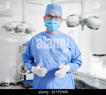 Porträt eines Arztes in einer Brille, der die Kamera anschaut, während er im Operationssaal steht. Mann Chirurg in sterilen Handschuhen, blaue chirurgische Uniform und schützende Gesichtsmaske, bereit für die plastische Chirurgie in der Klinik Stockfoto